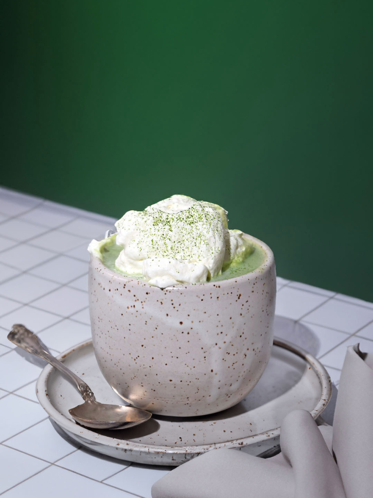 A coconut cream matcha in a speckled ceramic mug, topped with whipped cream and matcha powder, resting on a plate with a vintage spoon against a green background.