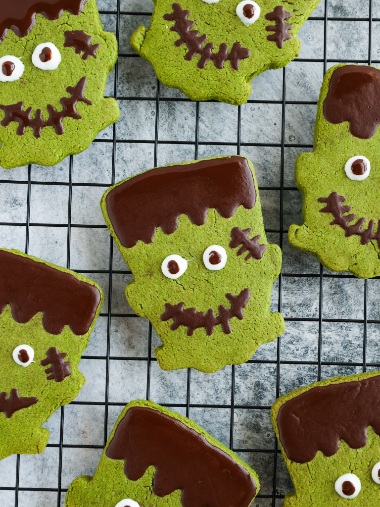 Homemade Matcha Frankenstein cookies on a cooling rack. Each cookie is shaped like Frankenstein’s head, vibrant green in color with dark chocolate hair and decorative scars. The cookies feature white and dark chocolate eyes