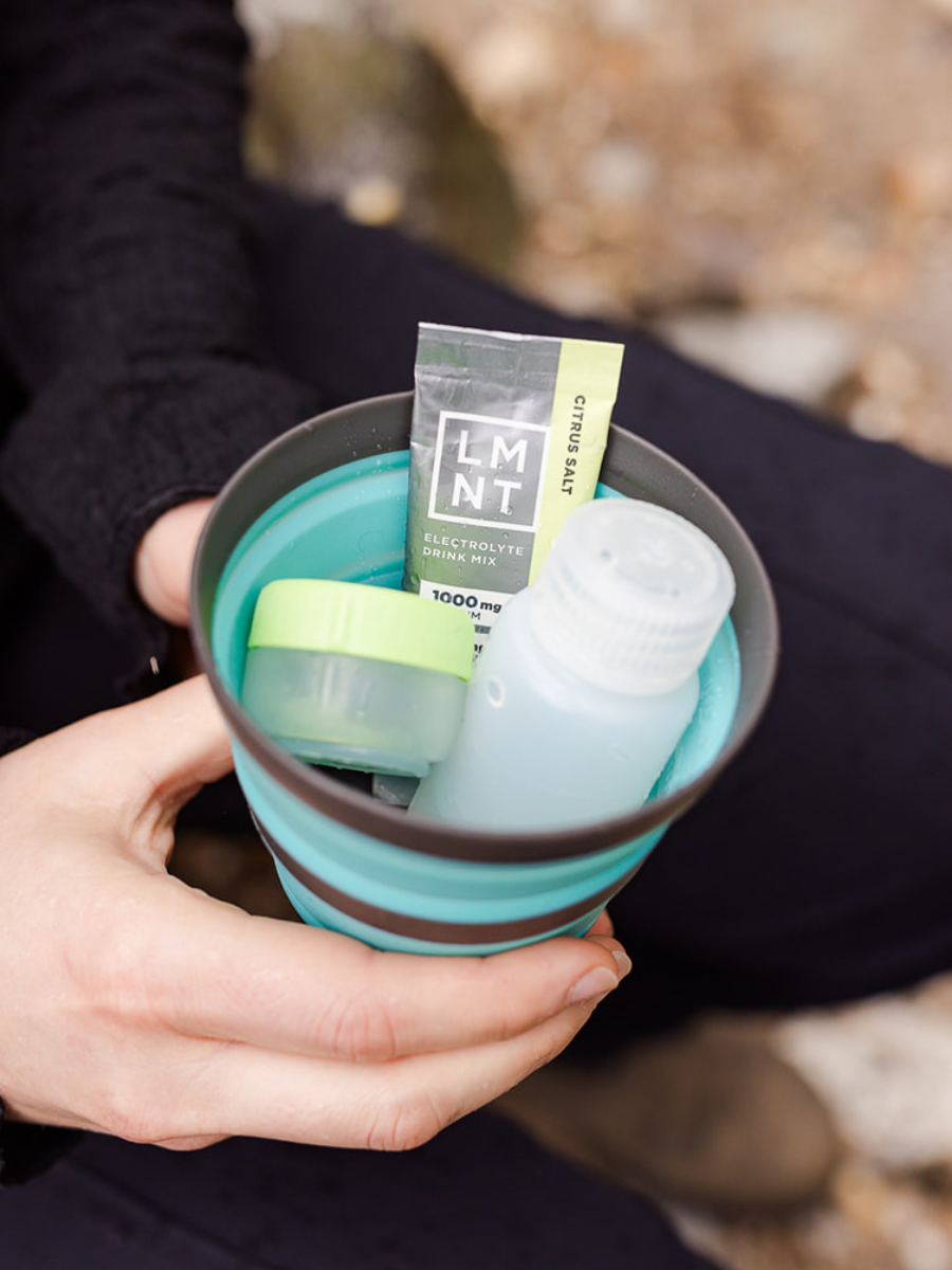 A person holding a collapsible turquoise travel cup filled with a packet of LMNT Citrus Salt electrolyte mix, a small container of matcha, and a mini bottle—perfect for on-the-go hydration during outdoor adventures.