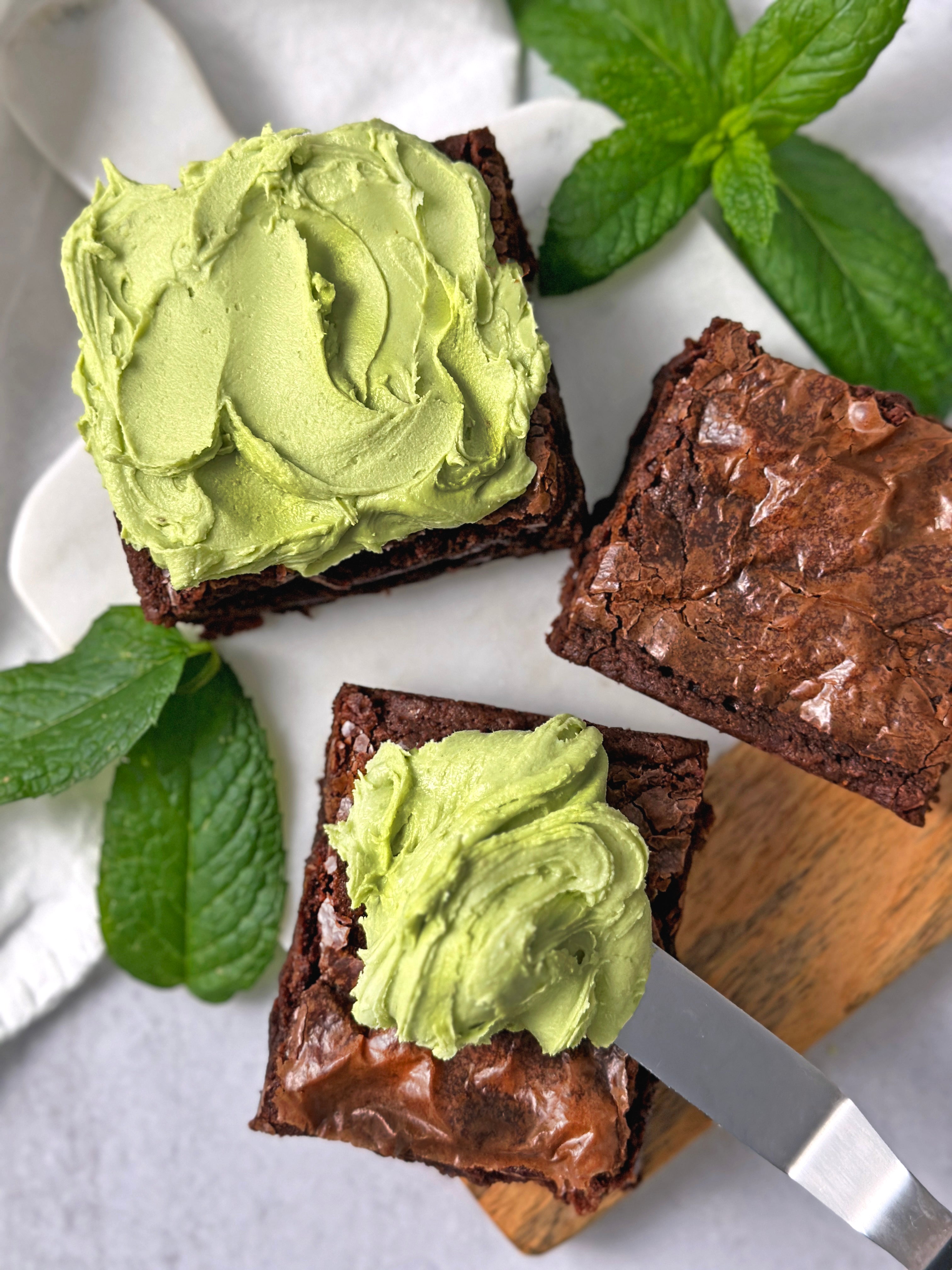 Rich chocolate brownies topped with vibrant green Matcha Mint Frosting, surrounded by fresh mint leaves.