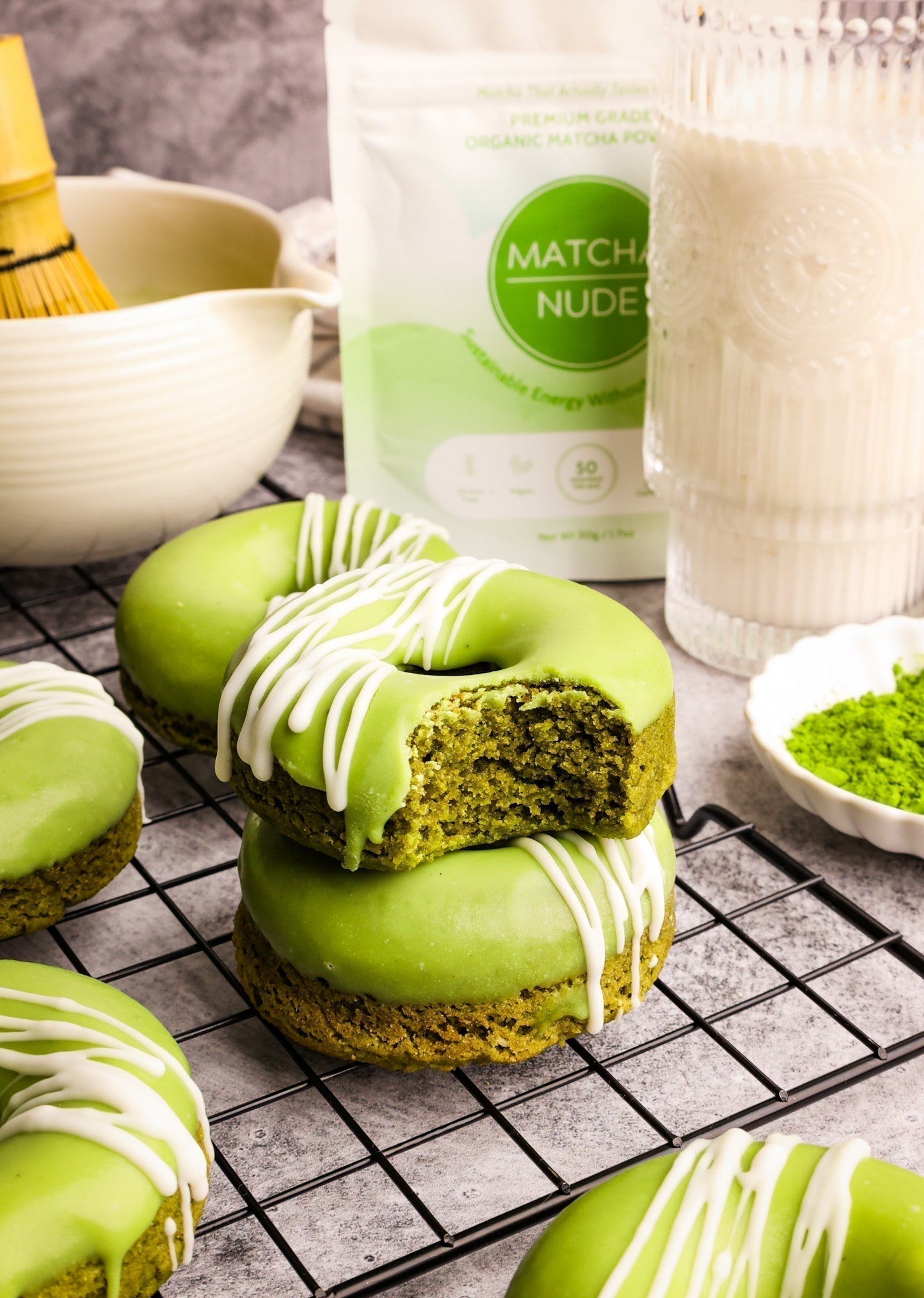  A stack of vibrant green matcha donuts with a drizzle of white icing sits on a cooling rack. The top donut has a bite taken out, revealing its fluffy texture. In the background, a bag of Matcha Nude organic matcha powder, a whisk, and a glass of milk.