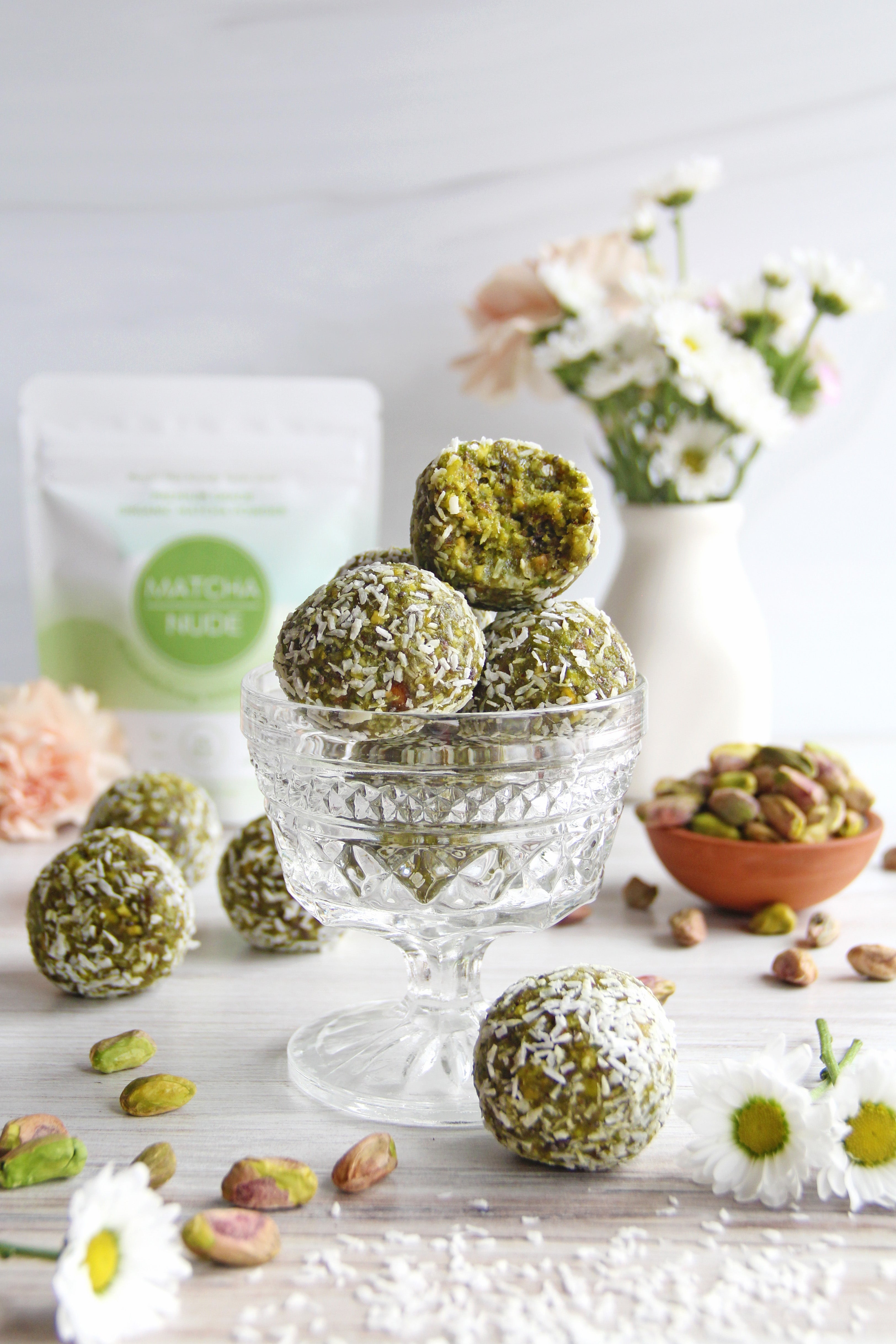 Matcha Pistachio Energy Bites coated in coconut, stacked in a glass dish with flowers and pistachios around.