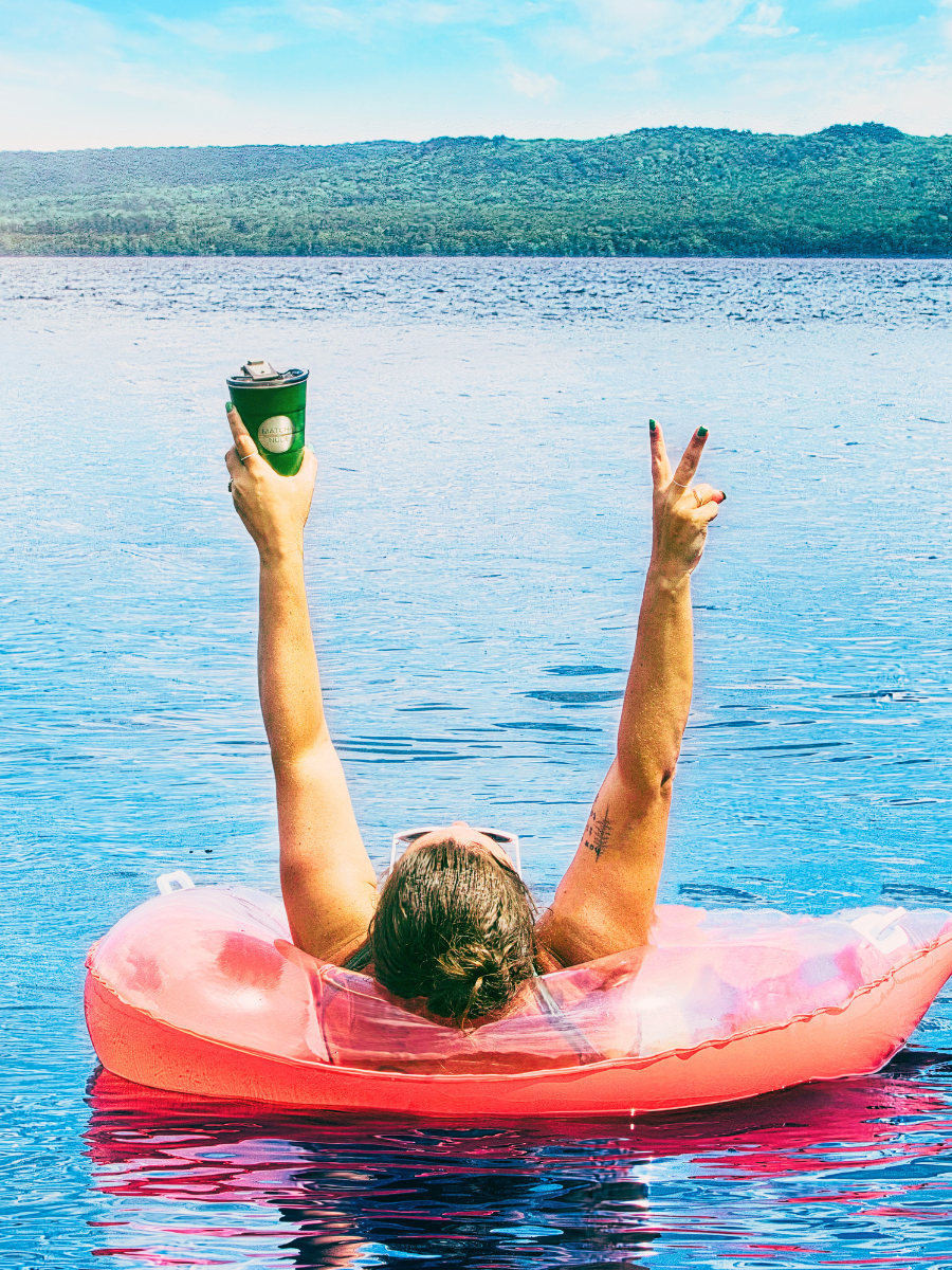 Person lounging on a pink inflatable tube in a serene lake, holding a green cup of matcha up in one hand and making a peace sign with the other. The sun is shining, and the landscape in the background features lush green hills under a clear blue sky.