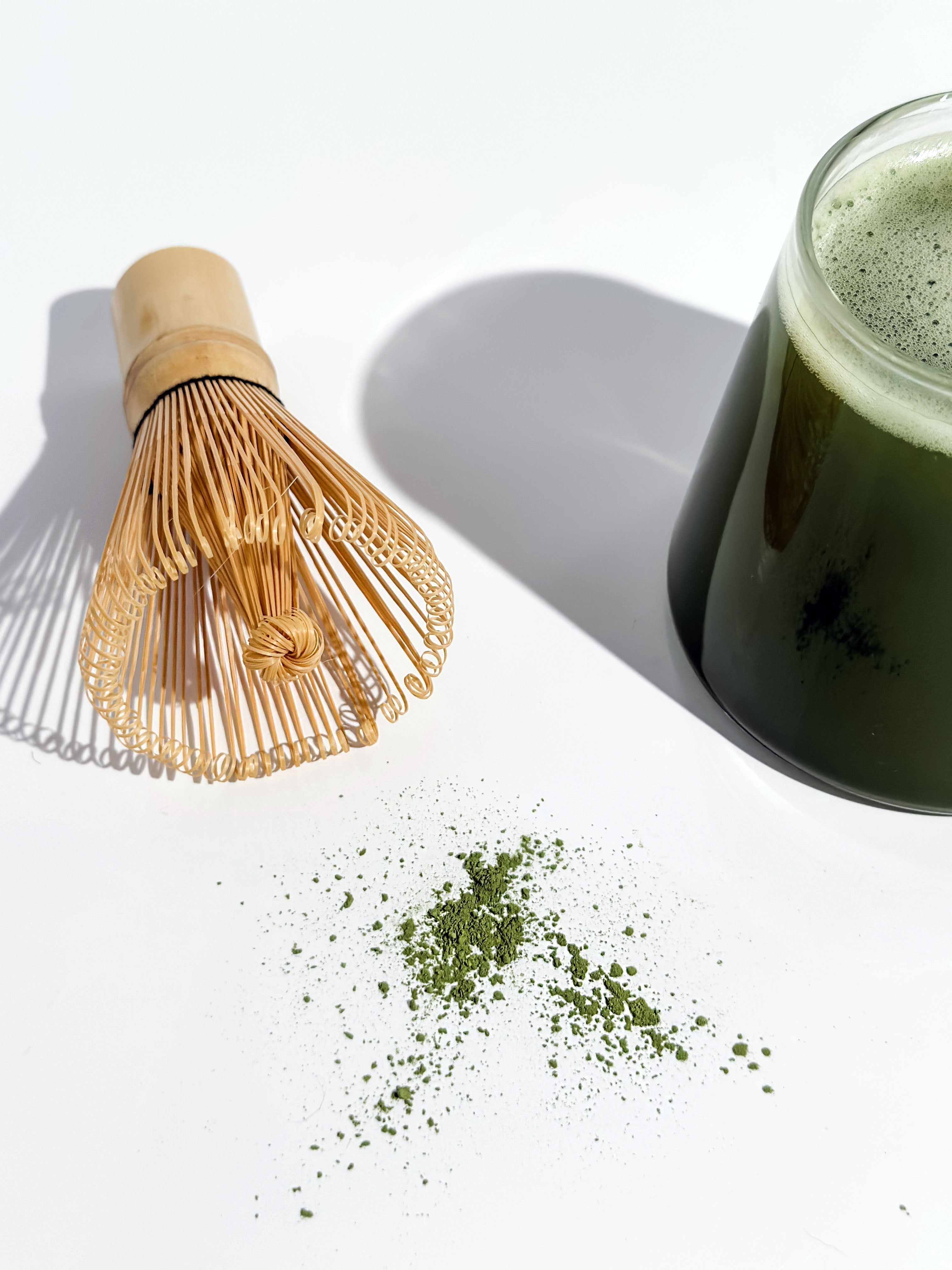 Loose matcha powder beside a bamboo whisk and glass of matcha.