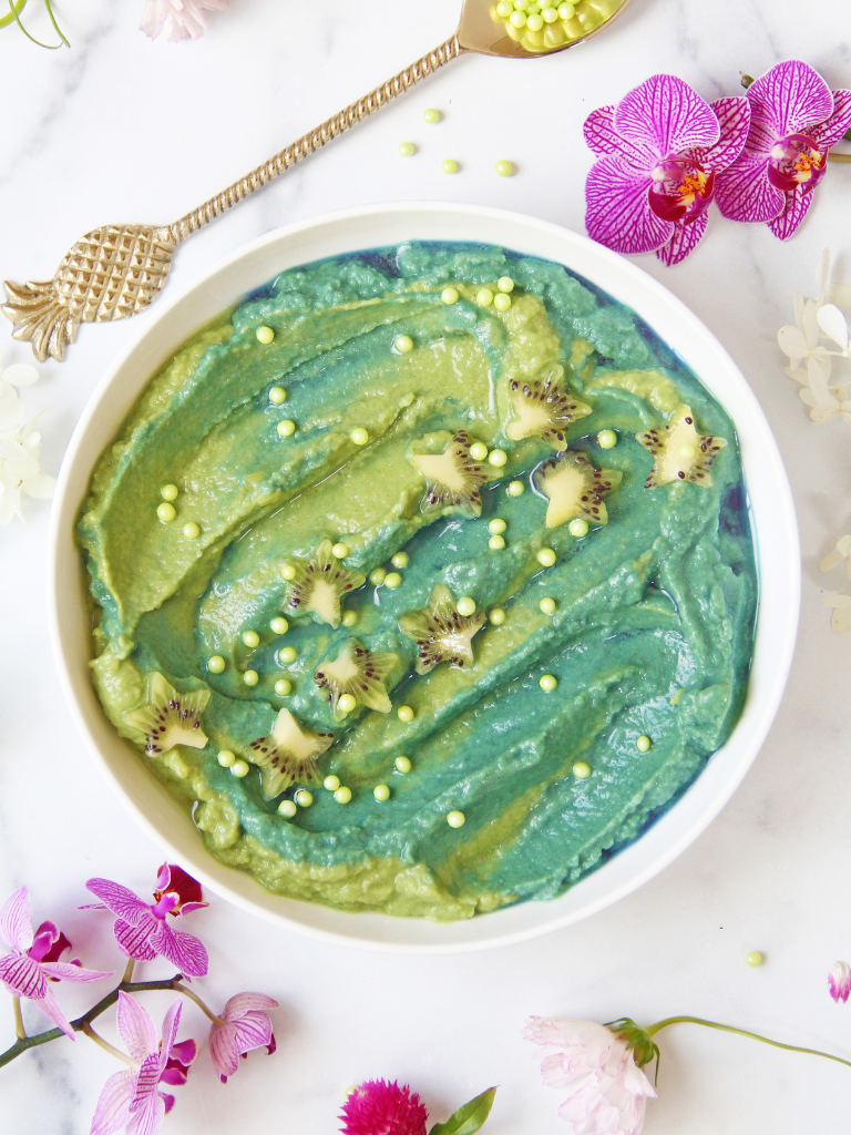 A vibrant bowl of Matcha Mermaid Pineapple Protein Nicecream, featuring swirling greens and blues, topped with kiwi and pearls. Served on a marble surface with a gold spoon and pink orchids, creating a whimsical, tropical look.