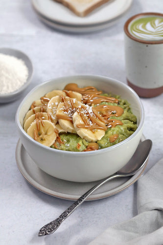 A cozy bowl of matcha oatmeal topped with sliced bananas, a drizzle of caramel, and a sprinkle of shredded coconut, served alongside a beautifully frothed matcha latte—perfect for a nourishing and delicious start to the day. 🍵✨
