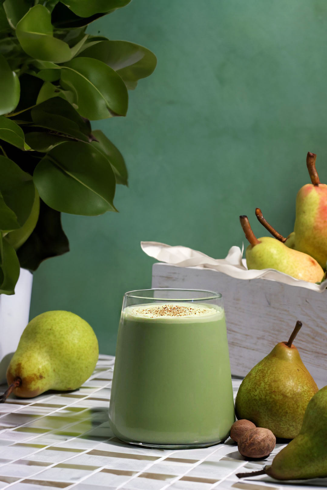 A glass of smooth, green matcha drink sprinkled with spices, surrounded by fresh pears and nutmeg on a tiled counter, next to a potted plant.