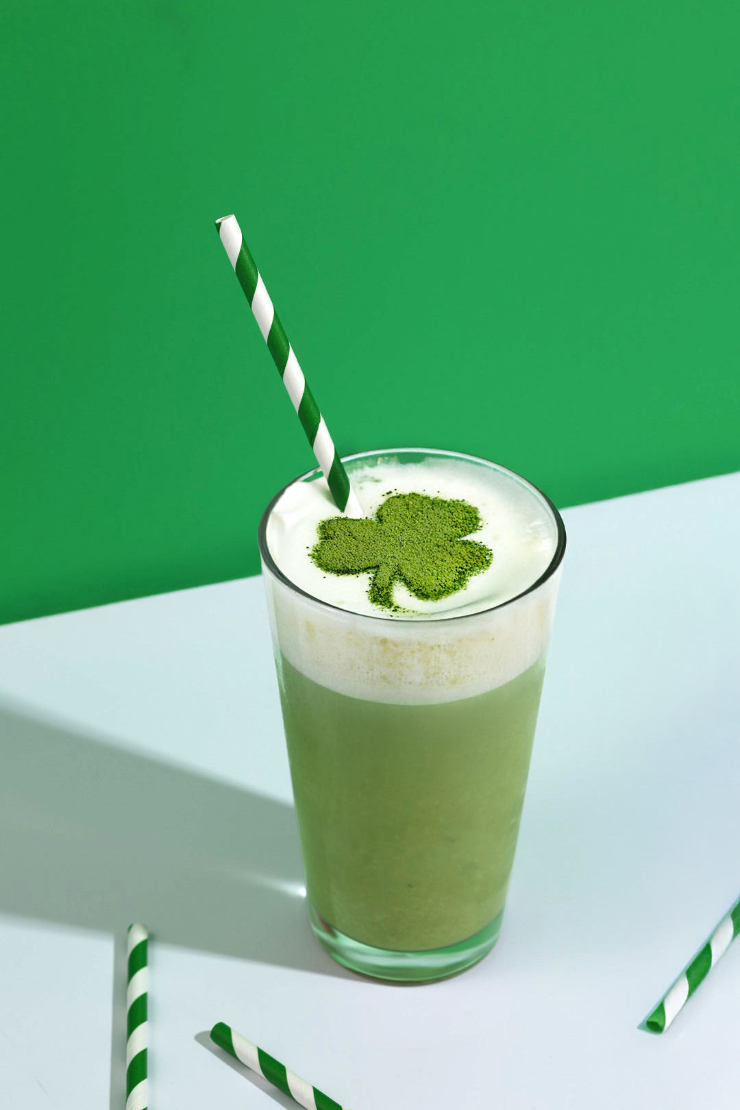 Shamrock Shake in a glass with a green and white striped straw and a shamrock shape made out of matcha powder on the top of the drink