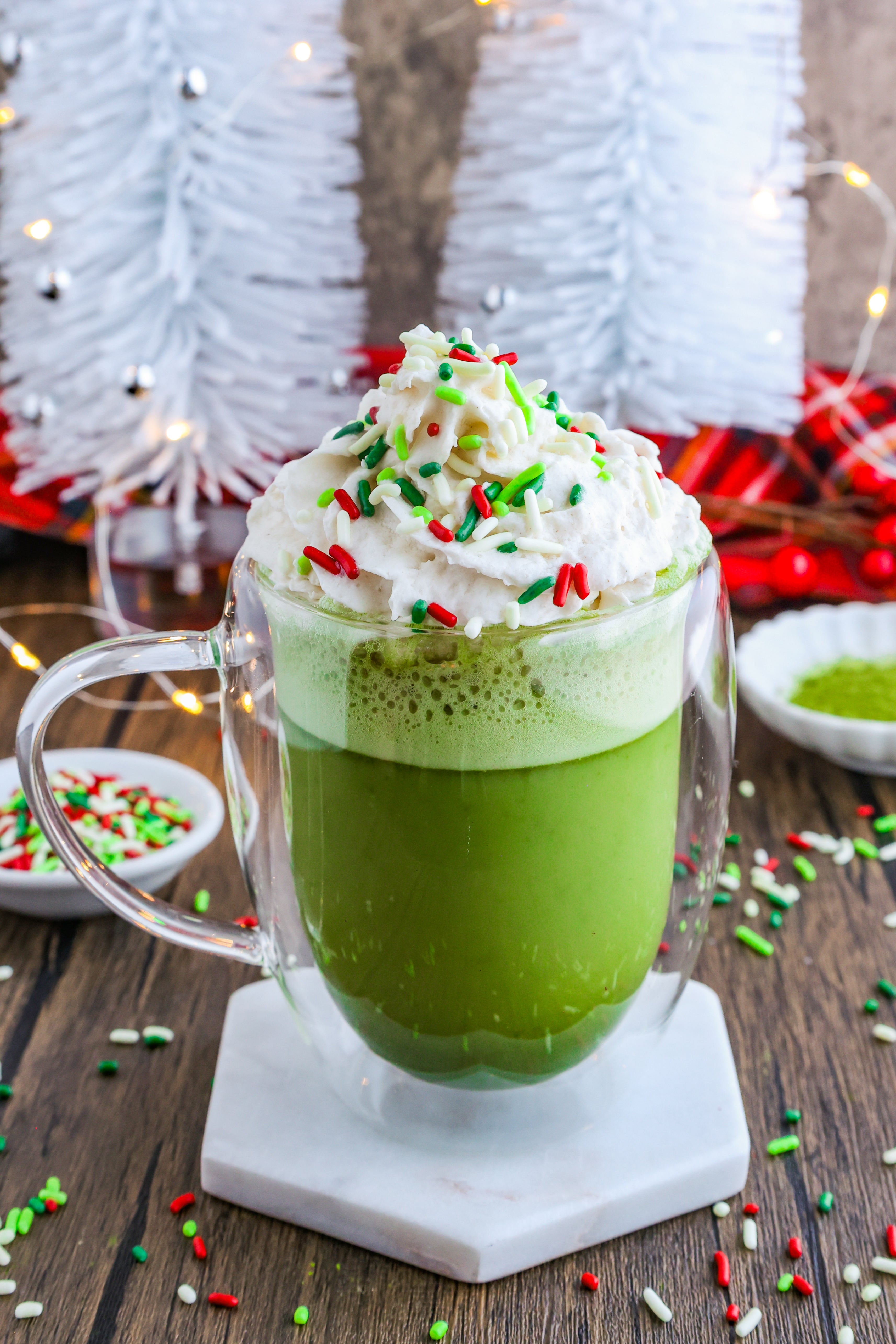 A festive Sugar Cookie Matcha Latte in a clear glass mug topped with fluffy dairy-free whipped cream and colorful red, green, and white sprinkles.