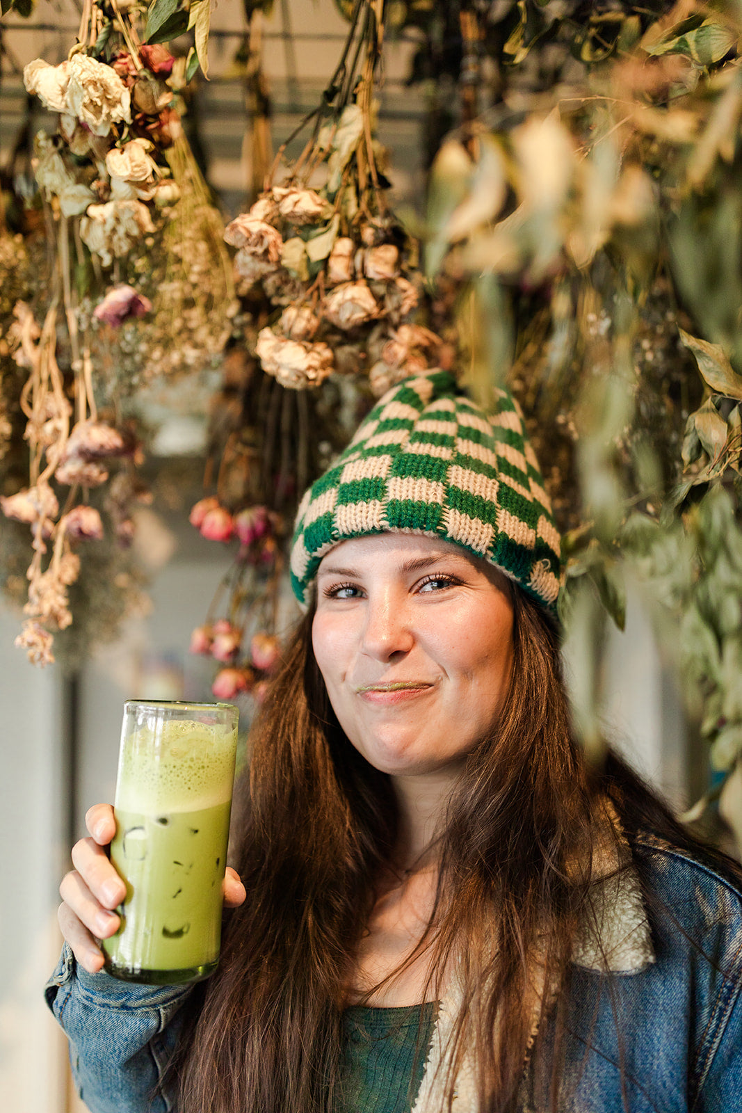 Woman in a green checkered beanie holding an iced matcha latte beneath hanging dried flowers.