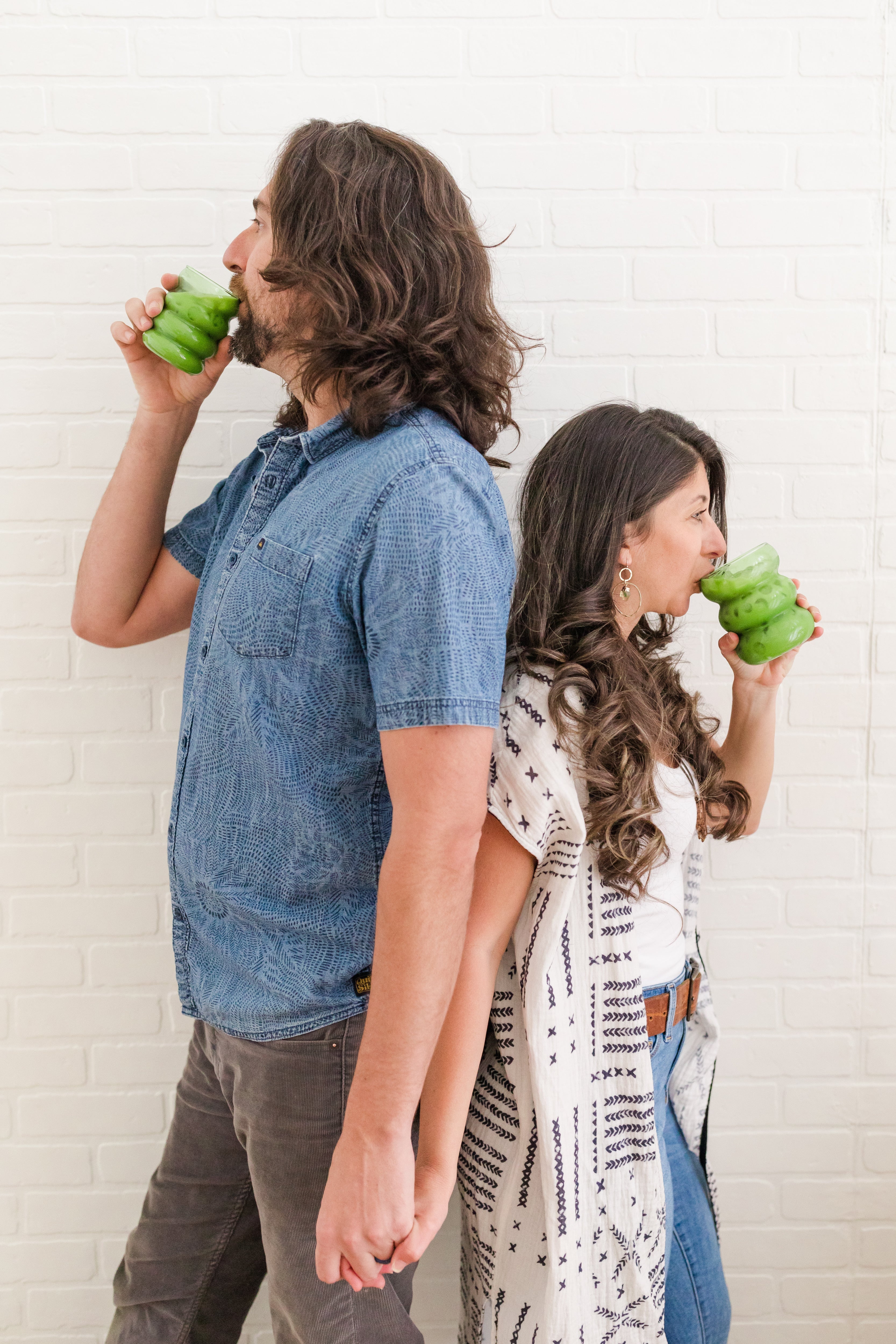 Man and woman standing back-to-back, sipping bright green matcha drinks against a white brick wall.