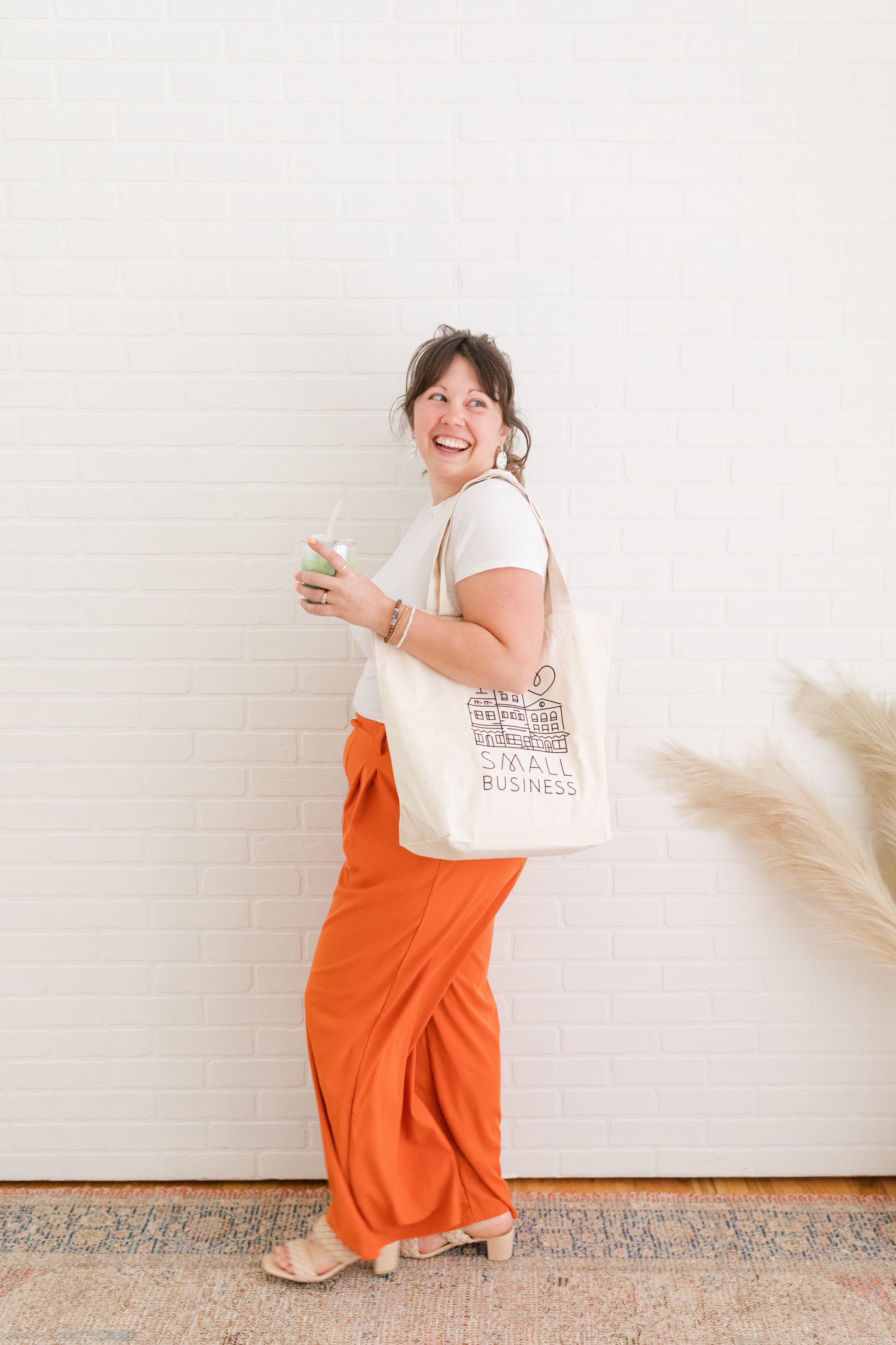 A woman holding an iced matcha drink and a tote bag, smiling as she stands against a white brick wall.