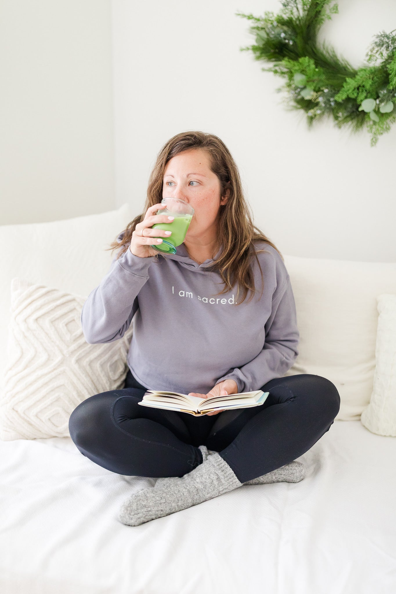 A woman sits on a cozy white couch, sipping a vibrant green matcha drink while holding an open journal. She’s wearing a lavender hoodie with “I am sacred” printed on it, exuding calm and focus. A lush green wreath hangs softly in the background.