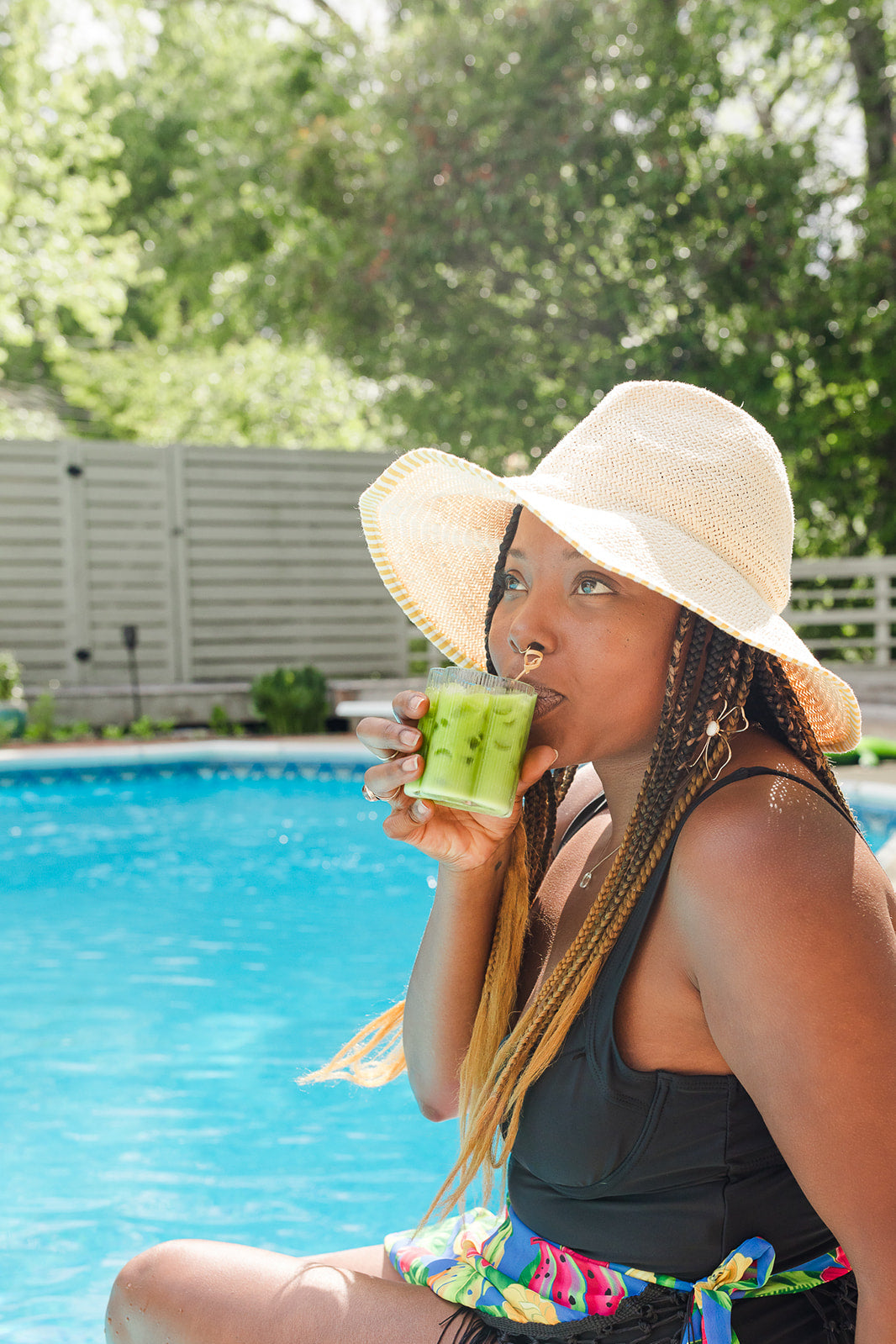 Woman sitting poolside wearing a sun hat, sipping an iced green matcha drink in bright sunlight.