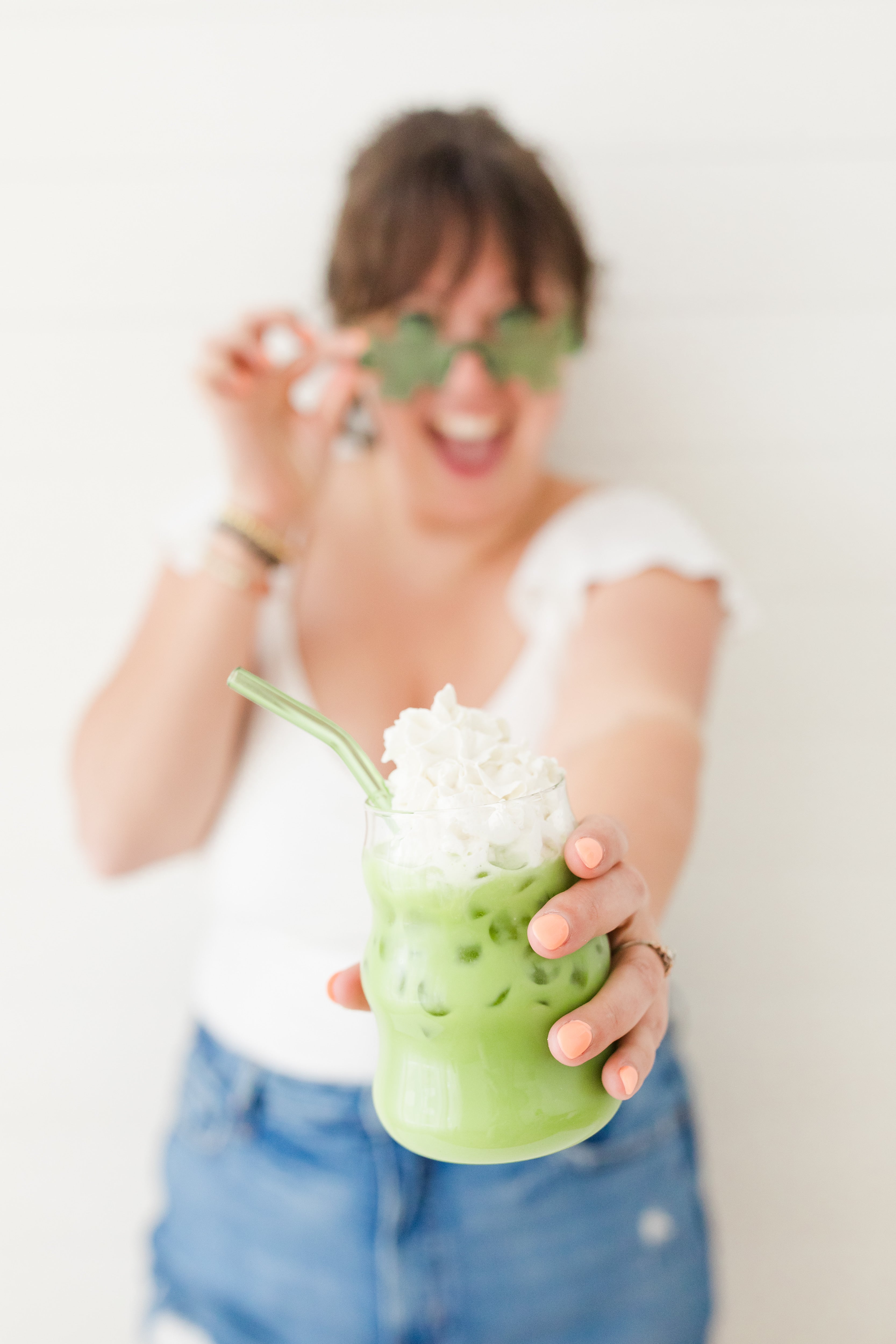 Person wearing shamrock-shaped sunglasses, smiling while holding an iced matcha latte with whipped cream and a green straw.