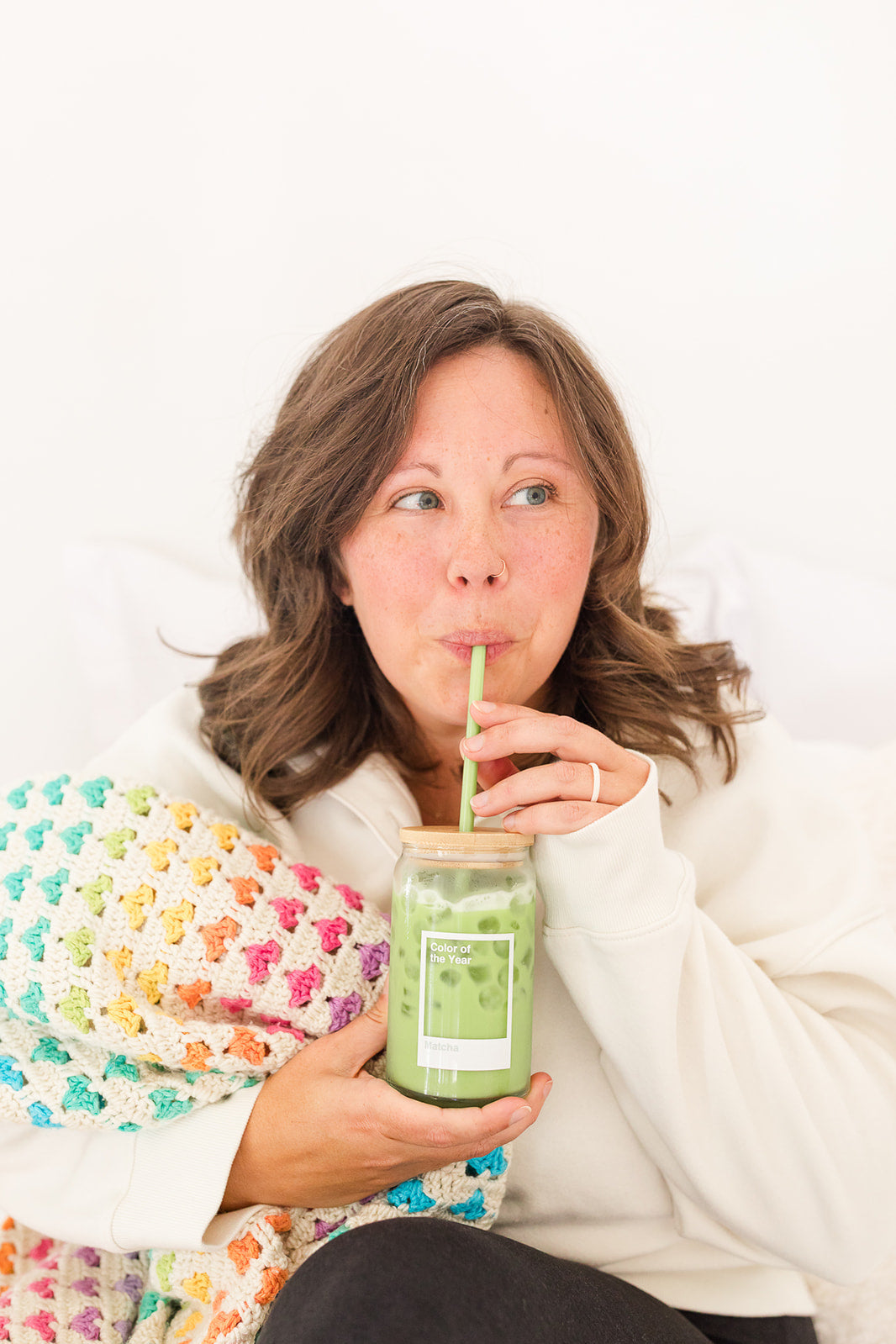 Woman sipping iced matcha latte from a branded glass jar while wrapped in a rainbow crochet blanket, representing Matcha Nude’s cozy, community-driven approach to clean energy and calm.