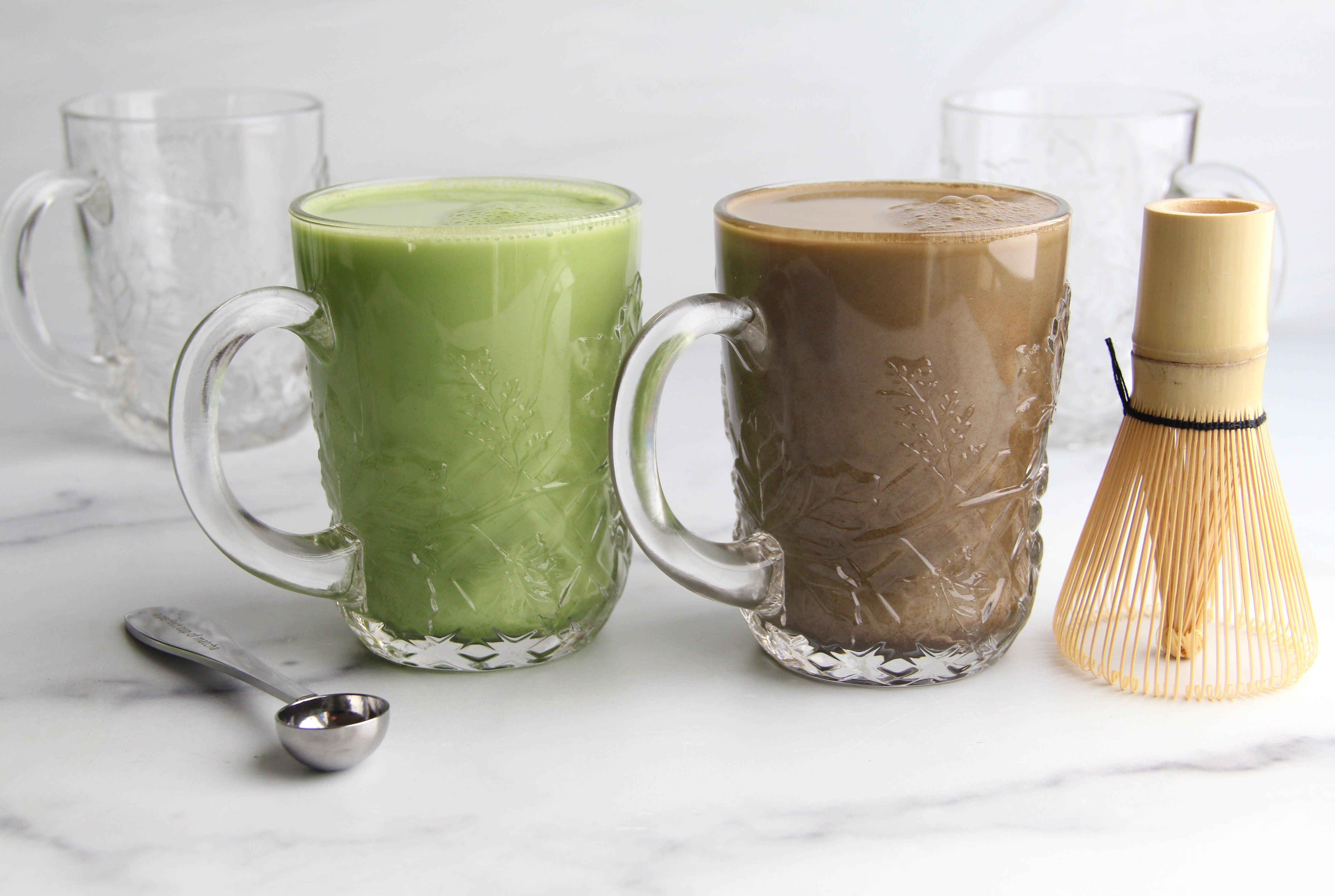 Two glass mugs of tea on a white marble surface, one bright green matcha latte and one rich brown hojicha latte next to a bamboo whisk and teaspoon.