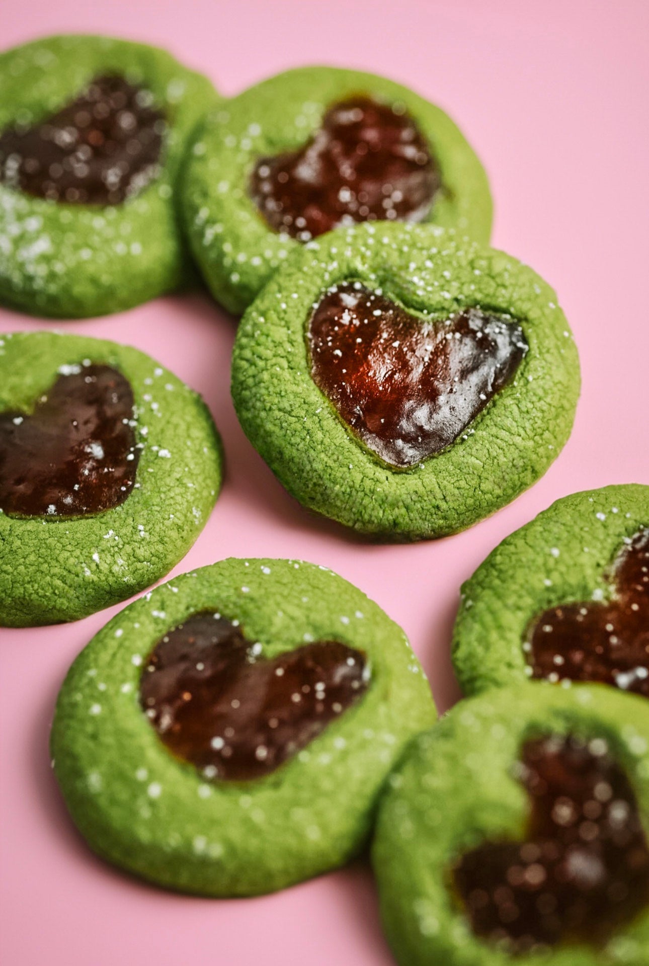 Heart-shaped Matcha Thumbprint Cookie filled with strawberry jam, dusted with powdered sugar on a pink counter.