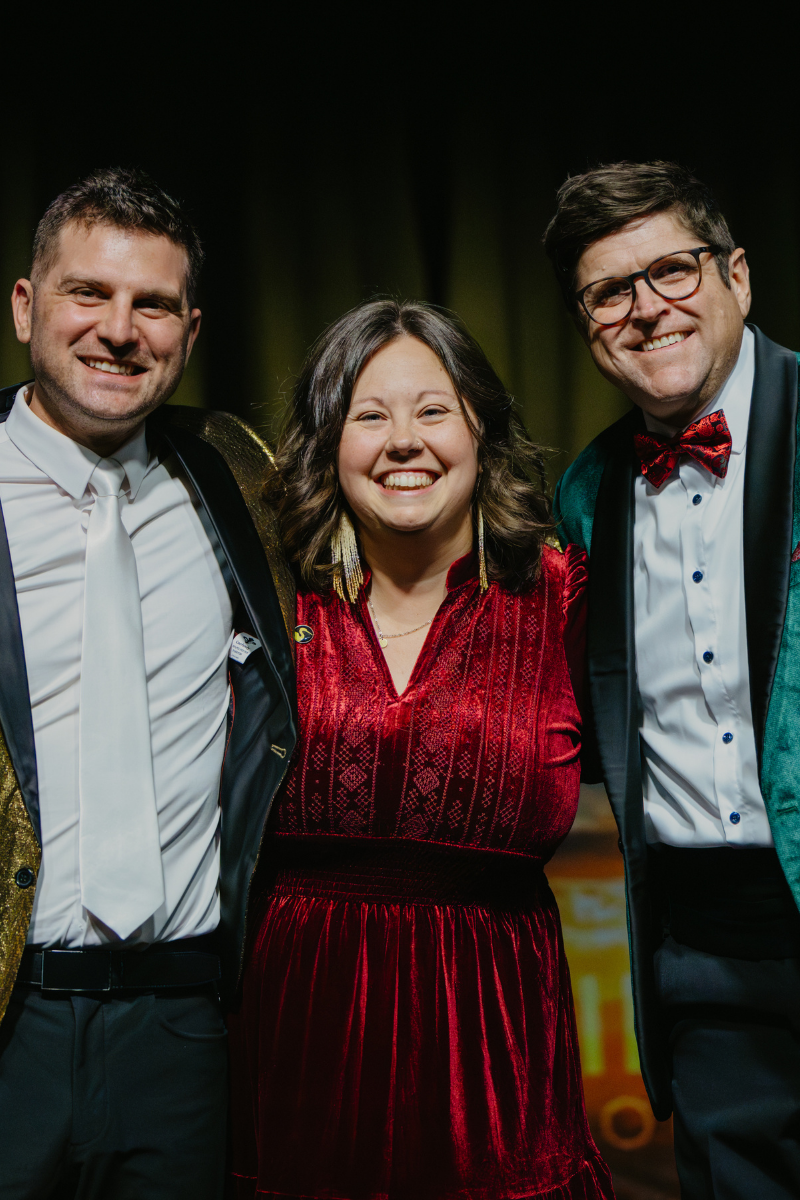Samantha Coffin standing onstage between two Venture Asheville hosts, smiling together; one wearing a gold blazer and the other a green velvet blazer.