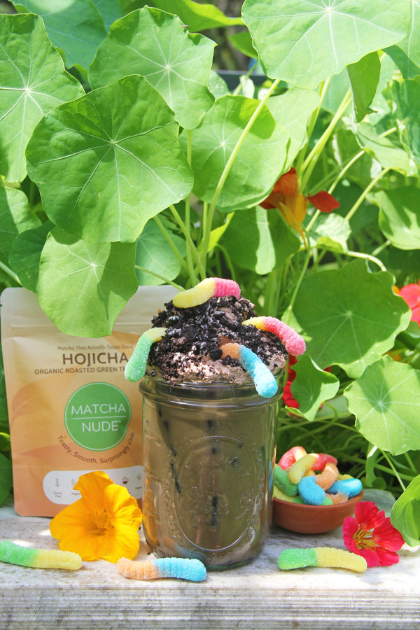 Hojicha Dirt latte topped with cookies and cream cold foam, cookie crumbles, and gummy worms styled with Matcha Nude Hojicha outdoors with plants.