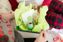 Load image into Gallery viewer, A gift box filled with Matcha Nude products, including a pouch of organic matcha powder, a bamboo whisk, a whisk stand, and a measuring spoon. The items are nestled in lime green tissue paper, creating a festive and vibrant display. The box is being held by a person dressed in cozy, holiday-themed pajamas.