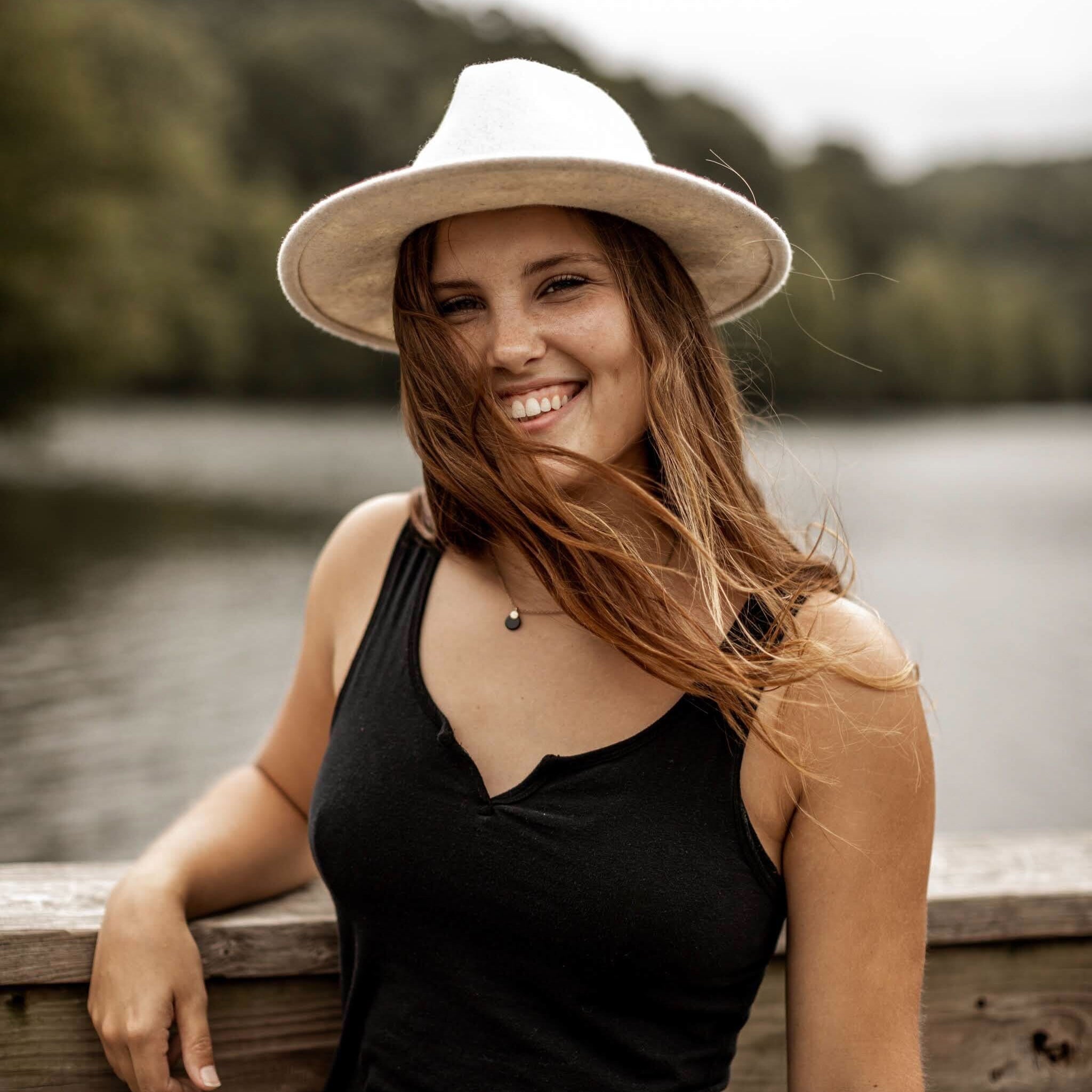 Smiling woman wearing a beige hat and black tank top standing by a lake with trees in the background.