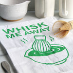 White tea towel with green “Whisk Me Away” text and matcha bowl illustration, styled with matcha whisk, mugs, and kitchen accessories on a countertop