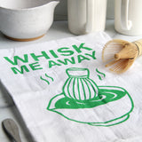 White tea towel with green “Whisk Me Away” text and matcha bowl illustration, styled with matcha whisk, mugs, and kitchen accessories on a countertop