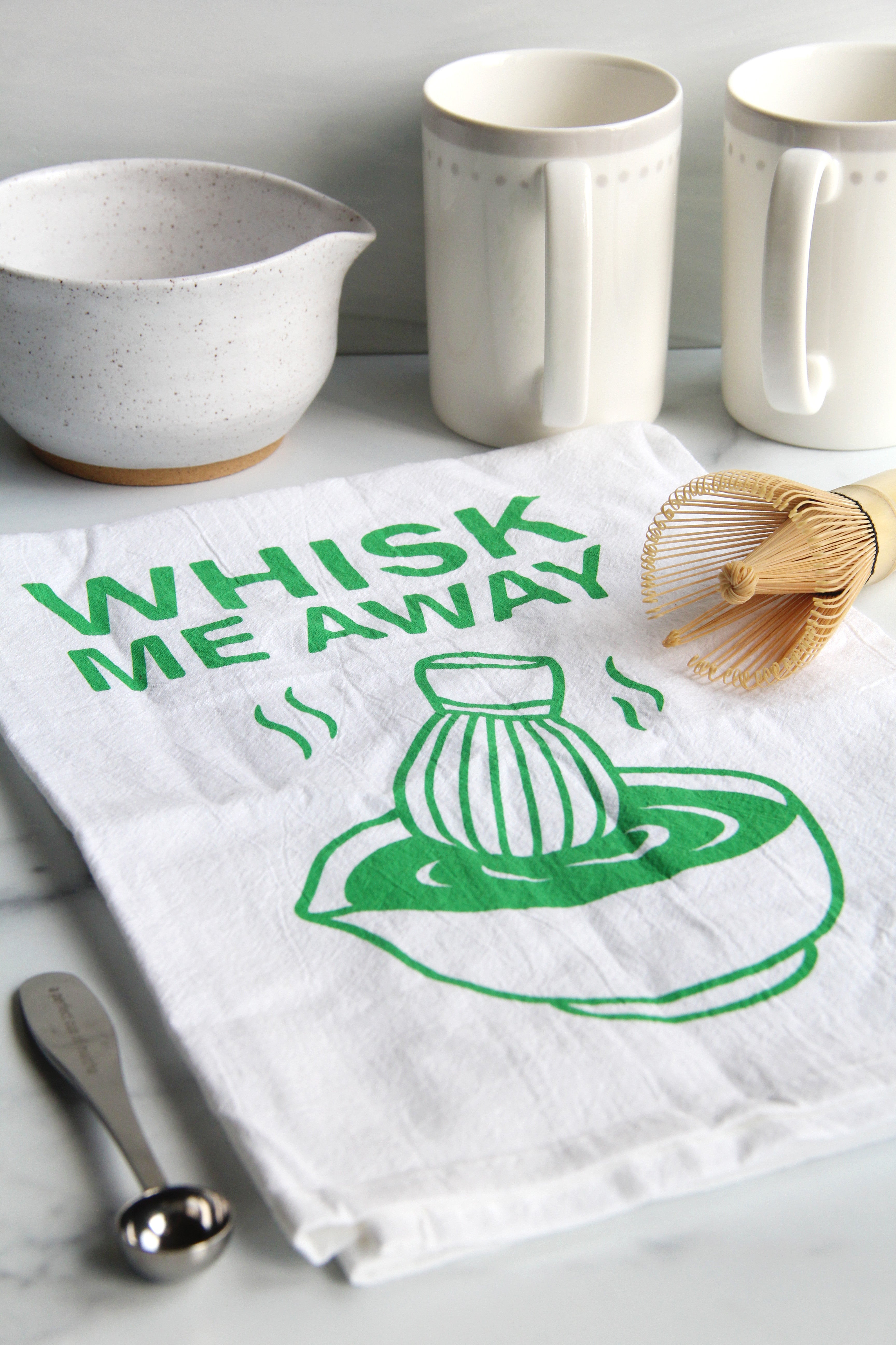 White tea towel with green “Whisk Me Away” text and matcha bowl illustration, styled with matcha whisk, mugs, and kitchen accessories on a countertop
