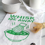 White tea towel with green “Whisk Me Away” text and matcha bowl illustration, styled with matcha whisk, mugs, and kitchen accessories on a countertop