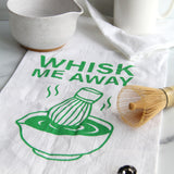 White tea towel with green “Whisk Me Away” text and matcha bowl illustration, styled with matcha whisk, mugs, and kitchen accessories on a countertop