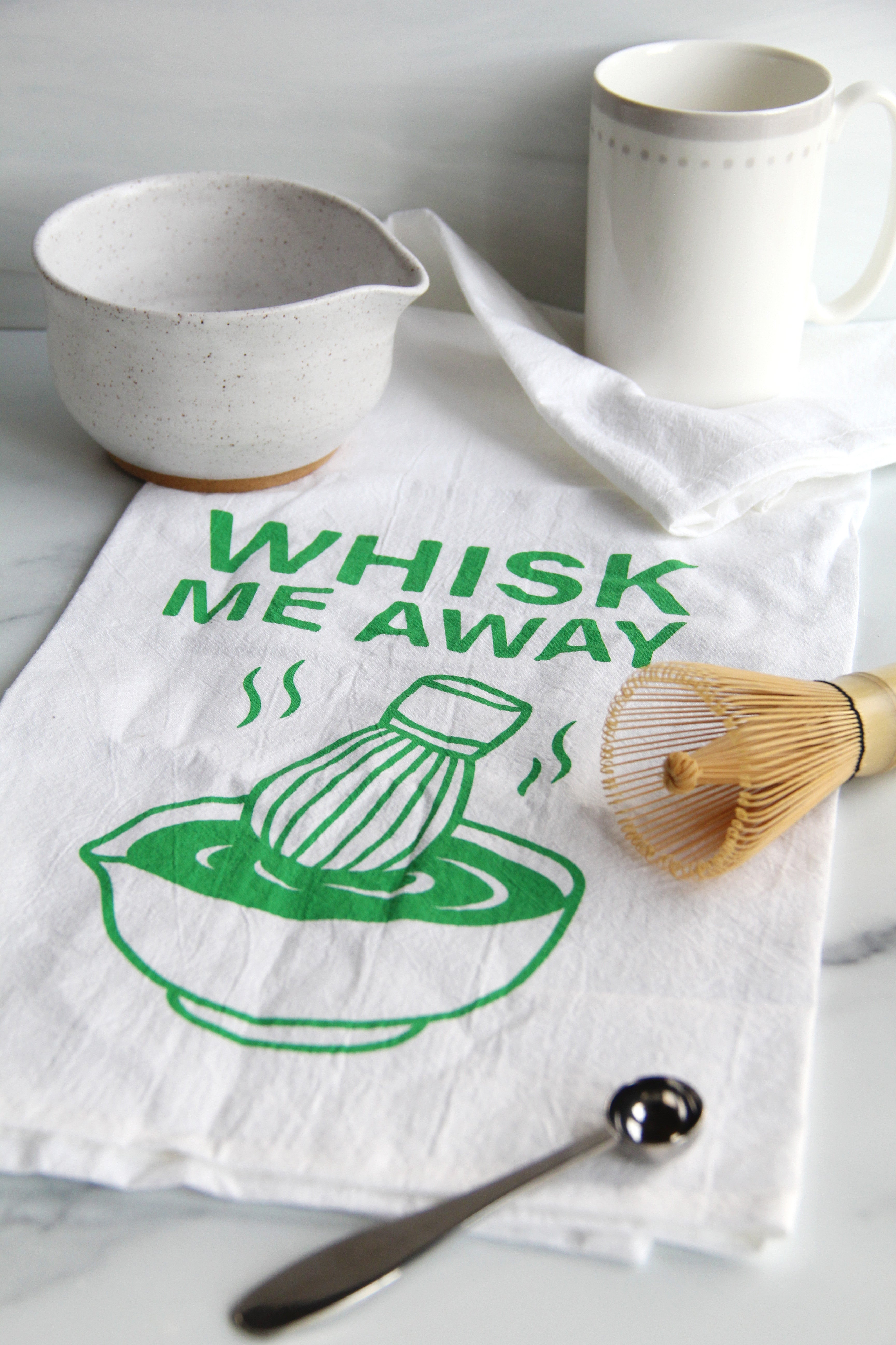 White tea towel with green “Whisk Me Away” text and matcha bowl illustration, styled with matcha whisk, mugs, and kitchen accessories on a countertop