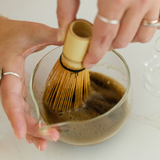 Person whisking Matcha Nude organic hojicha powder in glass bowl with bamboo whisk