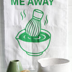 White tea towel with green “Whisk Me Away” text and matcha bowl illustration, styled with matcha whisk, mugs, and kitchen accessories on a countertop