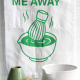 White tea towel with green “Whisk Me Away” text and matcha bowl illustration, styled with matcha whisk, mugs, and kitchen accessories on a countertop