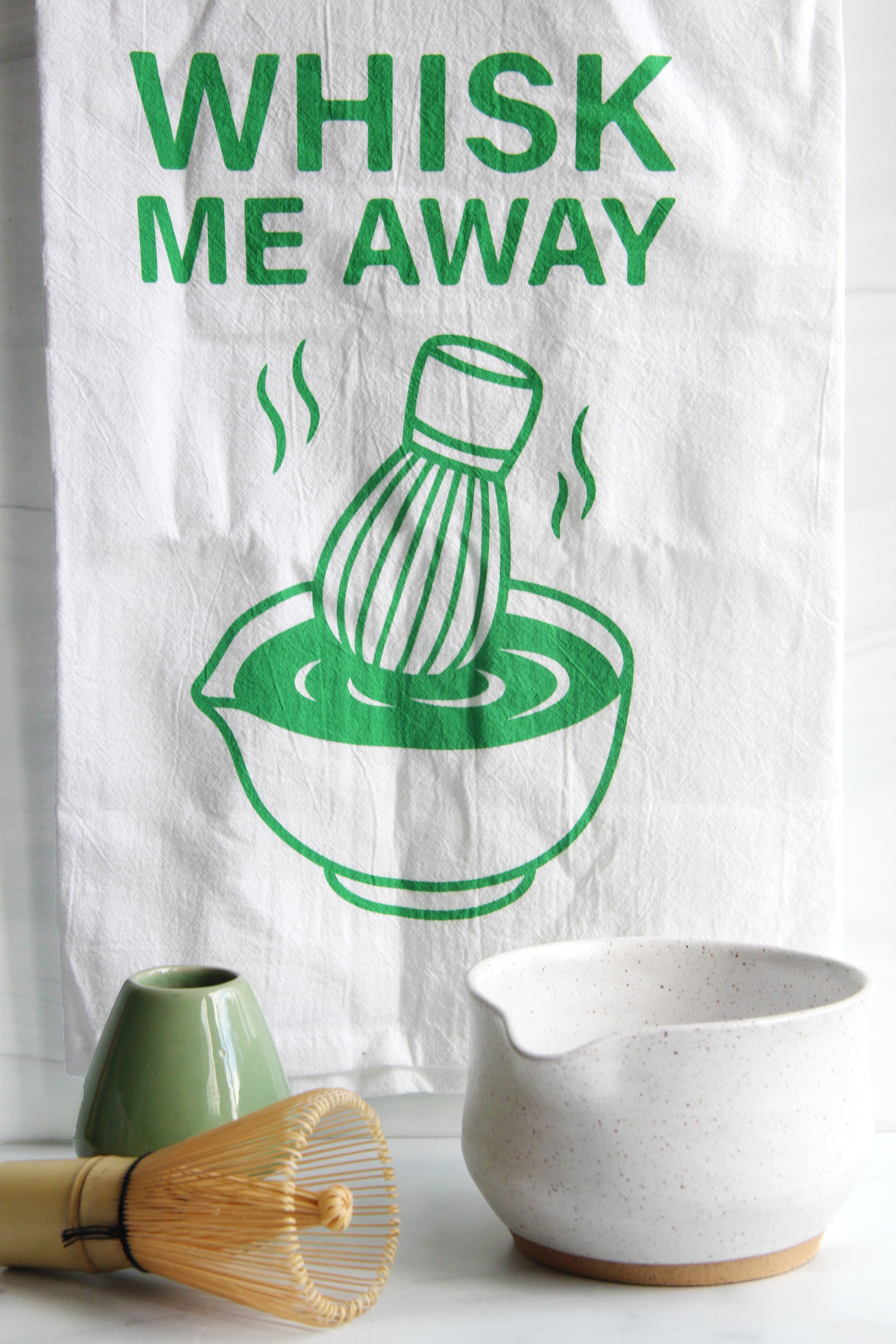 White tea towel with green “Whisk Me Away” text and matcha bowl illustration, styled with matcha whisk, mugs, and kitchen accessories on a countertop